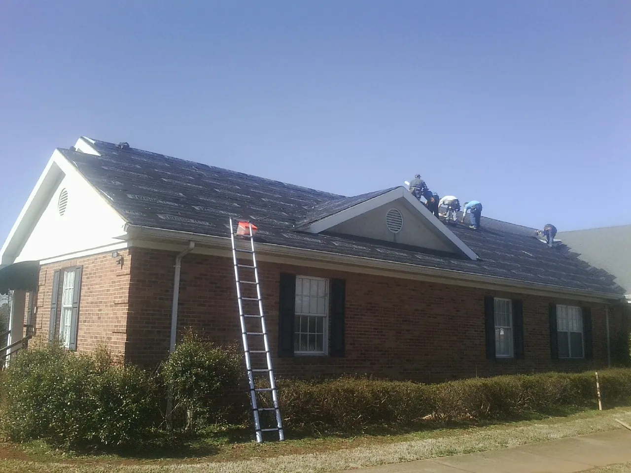 Crew tearing shingles off a brick ranch roof with a ladder set — Emergency Roof Repair work by our crew in Port Saint Lucie